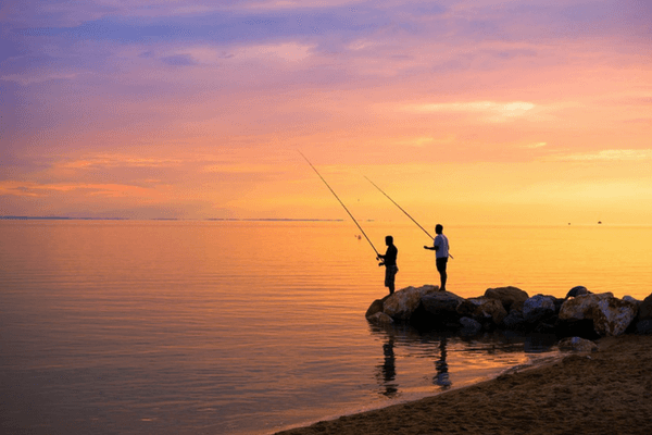 Two men standing on a rock and fishing in the sea during sunset